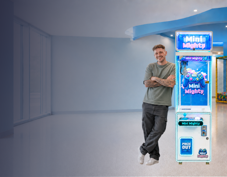 A man stands next to a tall blue and white vending machine in a brightly lit indoor play area or arcade with colorful play structures in the background.